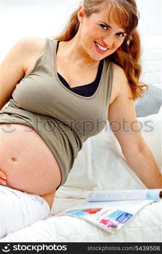 Smiling beautiful pregnant woman sitting on sofa at home with magazine.&#xA;