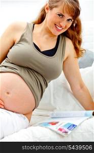 Smiling beautiful pregnant woman sitting on sofa at home with magazine.&#xA;