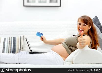 Smiling beautiful pregnant woman using credit card to shop from internet and showing thumbs up gesture&#xA;