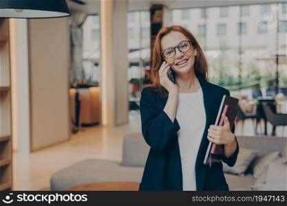 Smiling beautiful red-haired businesswoman manager making business call at work, happy female in eyewear consulting client by mobile while standing modern coworking space, holding laptop. Smiling beautiful red-haired businesswoman manager making business call at work
