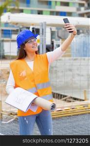 Smiling beautiful teenager wearing safety gear and holding blueprints takes a photo of herself outside an out of focus construction site. Work and apprenticeship concept.. Smiling teenager with safety gear takes a photo of herself