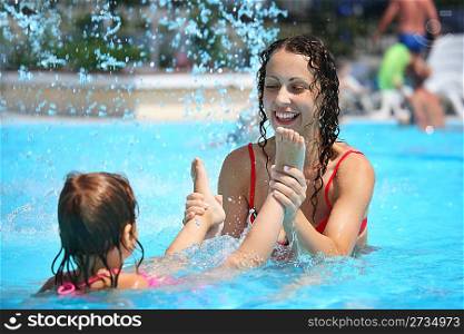 Smiling beautiful woman and little girl bathes in pool under water splashes, woman holds girl for feet