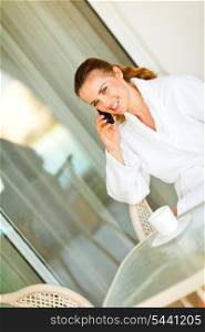 Smiling beautiful woman in bathrobe sitting at table on terrace and speaking phone&#xA;