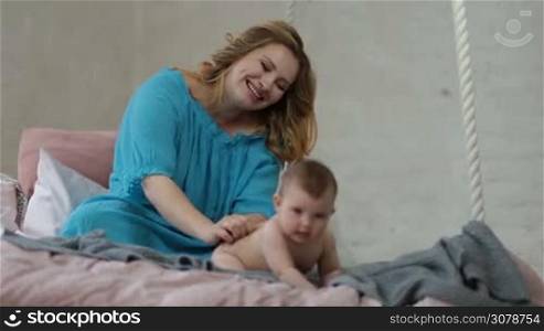 Smiling beautiful woman sitting on bed, playing, stroking and massaging naked infant child&acute;s back. Laughing little kid lying on her stomach naked on bed, enjoying massage by her affectionate mother in domestic bedroom.