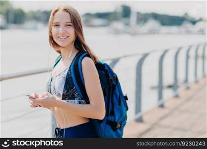 Smiling beautiful woman with happy expression, wears casual top, carries bag, holds cell phone in hands, enjoys online communication with friends, being always in touch, listens popular music