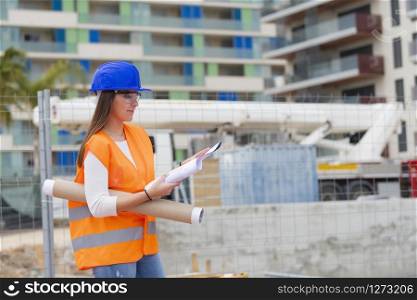 Smiling beautiful young architecture student wearing safety gear checking blueprints next to an out of focus construction site. Work and apprenticeship concept.. Architecture student checking blueprints next to a construction site