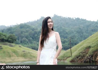 Smiling beautiful young Asian girl wear white dress standing on grass meadow beside the stream in tropical rainforest.