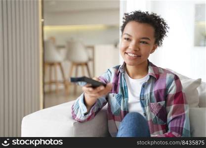 Smiling biracial american girl holding tv controller, switching channels, relax on couch at home, happy mixed race young woman spending leisure time with television, watching movie or series.. Happy biracial girl hold tv controller, watch movie or series, relax with television on sofa at home