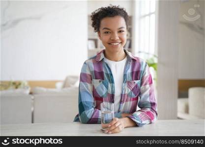 Smiling biracial girl with glass of fresh pure mineral water, sitting at table at home. Happy young female looking at camera enjoying purified water. Healthy lifestyle habit, self care concept.. Smiling biracial girl with glass of pure water, sitting at home. Healthy lifestyle habit, self care