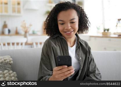 Smiling biracial young girl hold smartphone, chat with friend or post at social network, sitting on sofa at home. Friendly mixed race female using phone apps, shopping in online store on weekend.. Young biracial girl chat or post at social network, using smartphone apps, sitting on sofa at home