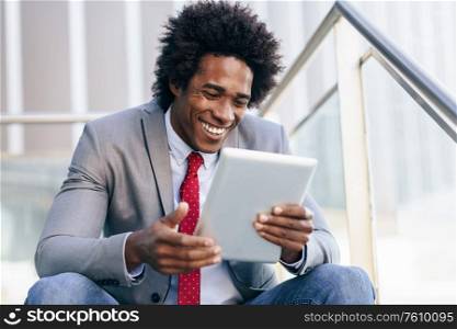 Smiling Black Businessman using a digital tablet sitting near an office building. Man with afro hair.. Black Businessman using a digital tablet sitting near an office building.