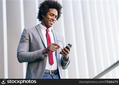 Smiling Black Businessman using his smartphone near an office building. Man with afro hair.. Black Businessman using a smartphone near an office building