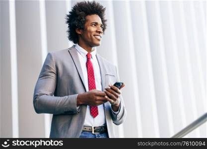 Smiling Black Businessman using his smartphone near an office building. Man with afro hair.. Black Businessman using a smartphone near an office building