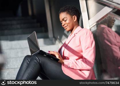 Smiling black businesswoman sitting on urban steps working with a laptop computer. African american female wearing suit with pink jacket.. Smiling black businesswoman sitting on urban steps working with a laptop computer