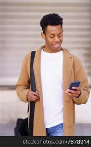 Smiling black man looking at his smartphone while walking down the street. Cuban guy in urban background.. Smiling black man looking at his smartphone while walking down the street.