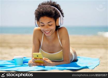 Smiling black woman lying on a towel on the sand on the beach using her smartphone to check Social Media. African American girl tanning.. Smiling black woman lying on the beach using her smartphone to check Social Media