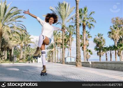 Smiling black woman on roller skates riding outdoors on beach promenade with palm trees. Smiling girl with afro hairstyle rollerblading on sunny day.