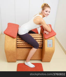 Smiling blond girl in sportwear sitting on modern relax massage equipment. Young woman doing relaxation exercise in healthy spa salon.