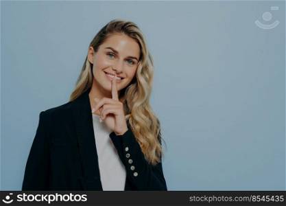 Smiling blonde girl in black coat keeping good news as secret, decides its better to wait before telling it or asking to keep silence while standing isolated on blue background. Secrecy concept. Smiling blonde girl keeping good news as secret