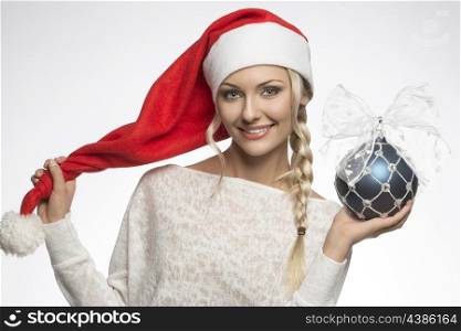 smiling blonde girl posing with white winter dress, red christmas hat and blue bauble, looking in camera