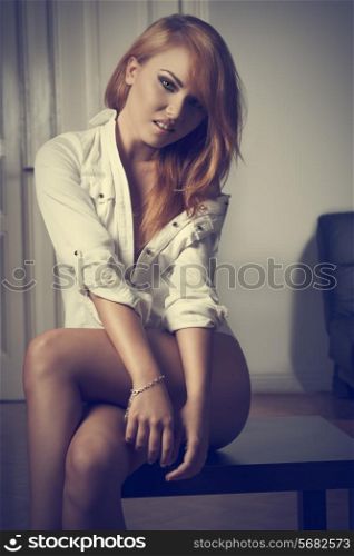 smiling blonde lady in erotic indoor portrait wearing only open shirt and showing her naked legs. Provocative pose