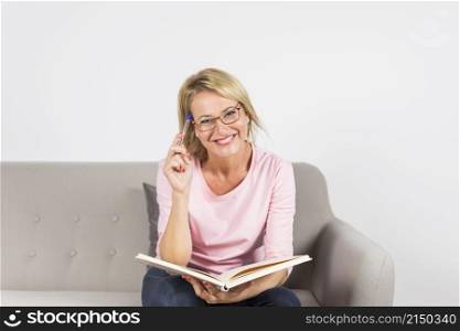 smiling blonde mature woman sitting sofa holding pen book against white backdrop