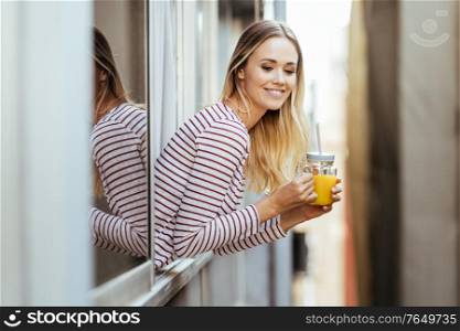 Smiling blonde woman drinking a glass of natural orange juice, leaning out the window of her home.. Smiling woman drinking a glass of natural orange juice, leaning out the window of her home.