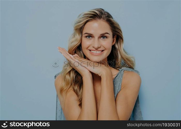 Smiling blonde woman in blue dress resting head on hands held together, looking straight at camera with happiness in her eyes while standing isolated next to blue background. Women beauty concept. Smiling blonde woman resting head on hands held together