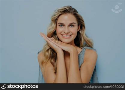 Smiling blonde woman in blue dress resting head on hands held together, looking straight at camera with happiness in her eyes while standing isolated next to blue background. Women beauty concept. Smiling blonde woman resting head on hands held together