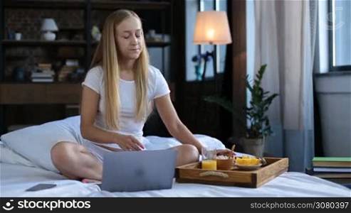 Smiling blonde woman in white underwear eating oatmeal while working on laptop in bed in the morning. Positive young female sitting crossed legs on white bed sheet socializing in networks using laptop pc during morning healthy breakfast in bedroom.