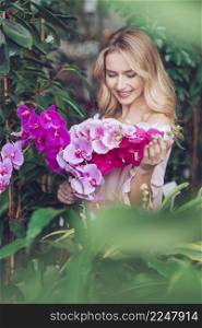 smiling blonde young woman standing front green plants looking exotic pink orchid flowers