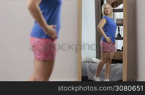 Smiling body-positive blonde woman in pajamas admiring her shape in the mirror at home. Charming cheerful girl looking at her reflection in the mirror and happy to see her slimmed figure.