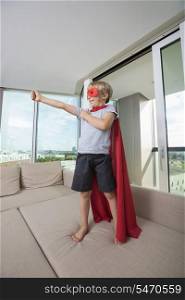 Smiling boy in super hero costume standing with arm outstretched on sofa bed at home