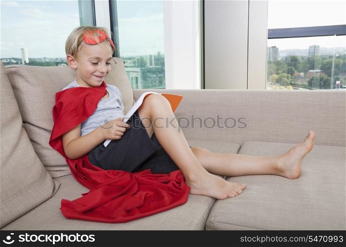 Smiling boy in superhero costume reading book on sofa at home
