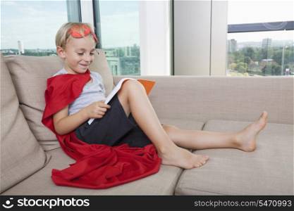 Smiling boy in superhero costume reading book on sofa at home
