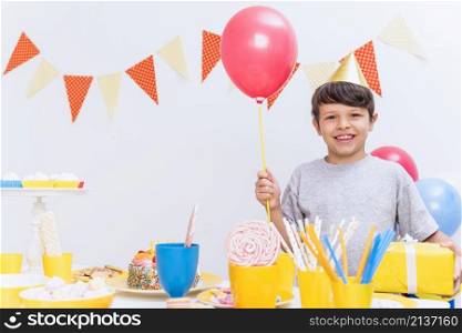 smiling boy wearing party hat holding balloon gift standing variety food table