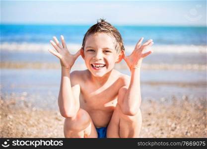 smiling boy with hands close to the head sitting on the beach. smiling boy sitting on the beach