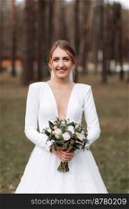 smiling bride in a long white dress is standing in the park and holding a wedding bouquet of roses flowers and greens. young woman with hairstyle, makeup and long veil outdoors near hotel.. smiling bride in a long white dress is standing in the park and holding a wedding bouquet of roses flowers and greens. young woman with hairstyle, makeup and long veil outdoors near hotel