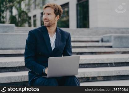 Smiling brown-haired neat unshaven office worker in stylish suit sitting outside on stairs in city center with laptop on his lap, checking important emails and reports from work. Remote job outdoors. Young smiling office worker sitting outside on stairs in city center holding laptop on his lap