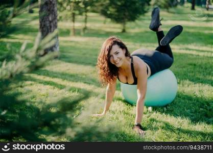 Smiling brunette exercises on green grass with fitness ball, aiming for a sporty body. Curly hair, sportswear, outdoor yoga training on a warm sunny day.