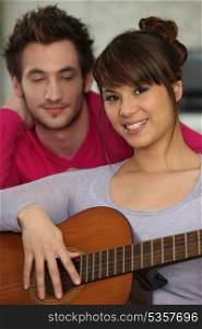smiling brunette playing the guitar under boyfriend&rsquo;s watchful eye