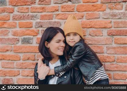 Smiling brunette woman and her little daughter embrace with love, have charming smiles, wears black leather jackets, pose against brick wall. Young brunette mother spends free time with female kid