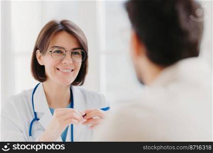 Smiling brunette woman doctor in round glasses, wears white coat, has conversation with patient, happy to help other people, wears white coat uniform, discuss health care. Good treatment concept