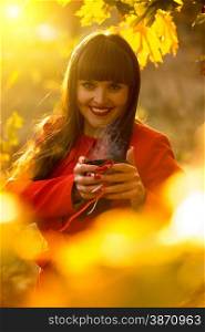 Smiling brunette woman holding evaporating cup of tea at autumn park