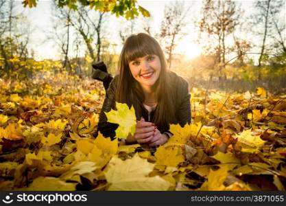 Smiling brunette woman lying on ground covered with leaves at autumn park