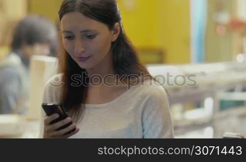 Smiling brunette woman types message using smartphone while sitting in cafe
