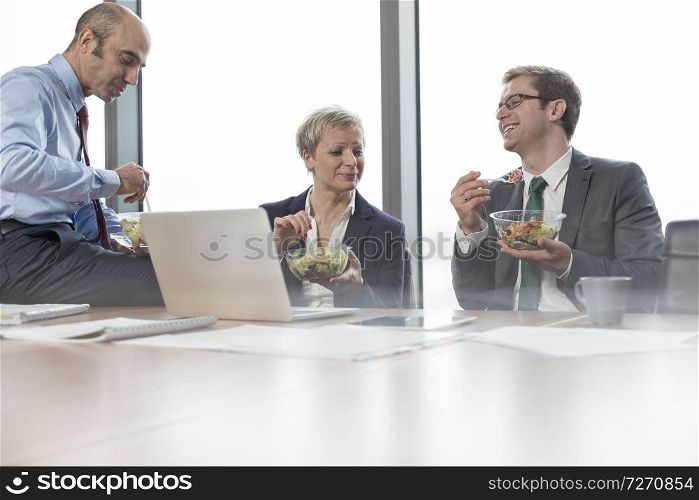 Smiling business colleagues eating lunch while sitting in boardroom during meeting at office