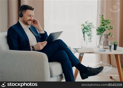 Smiling business person in suit wearing headset having video call, working online on laptop while sitting in living room in comfortable armchair. Business people and remote work at home. Happy male manager enjoying distant work at home