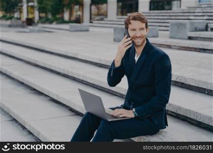 Smiling business professional in classic two piece suit speaks on smartphone with customers, young attractive businessman working on laptop computer distantly outside while sitting on stairs. Young attractive businessman working on laptop distantly, speaking on smartphone outside