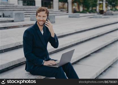 Smiling business professional in classic two piece suit speaks on smartphone with customers, young attractive businessman working on laptop computer distantly outside while sitting on stairs. Young attractive businessman working on laptop distantly, speaking on smartphone outside